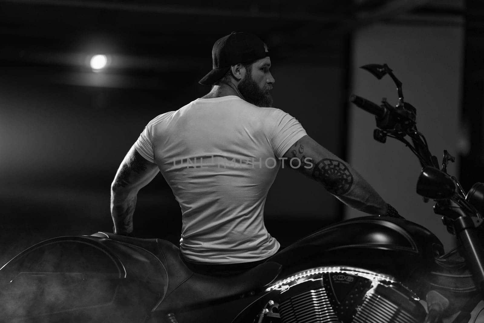 Sexy handsome attractive sporty muscular fitness model breaded biker with brutal beautiful motorcycle in the parking posing with a motorbike with his back to the frame, black and white photo