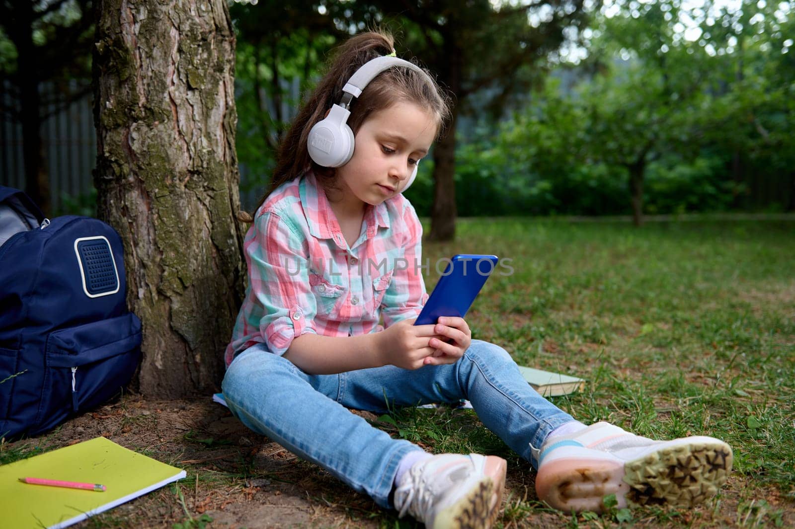 Cute little child girl, happy primary school student holding a smart mobile phone, listening to music on headphones, playing online video games, relaxing in the park after class, sitting on the grass