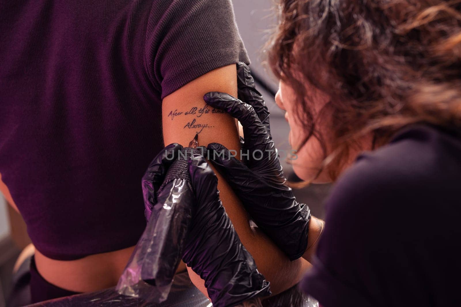 Talented focused woman tattoo artist diligently crafting personalized lettering tattoo using machine on arm of female client, cropped shot. Tattooing process concept