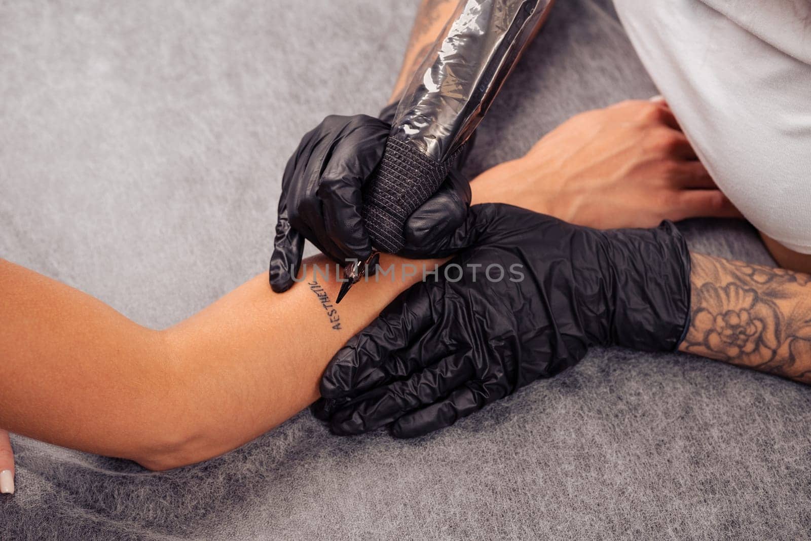 Hands of professional skillful master getting tattoo of word AESTHETIC on arm of female client, cropped shot. Concept of creating of individual body design