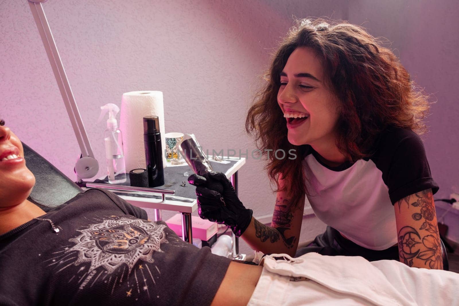 Cheerful young girl, professional tattoo artist, friendly chatting with client and laughing while getting tattoo in salon