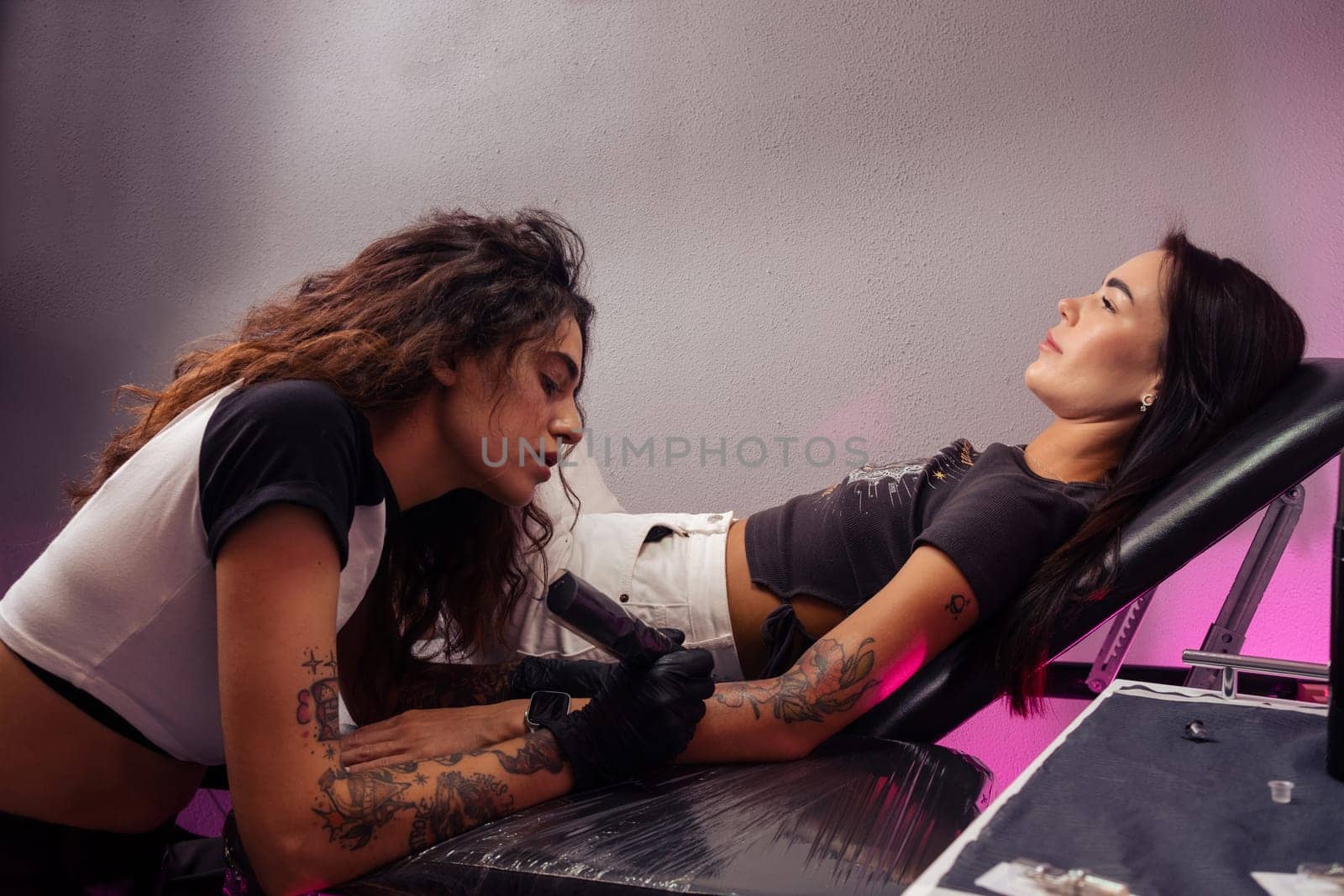 Skilled young female tattooist at work. Focused professional tattoo master creating colorful artistic masterpiece on female client arm in parlor