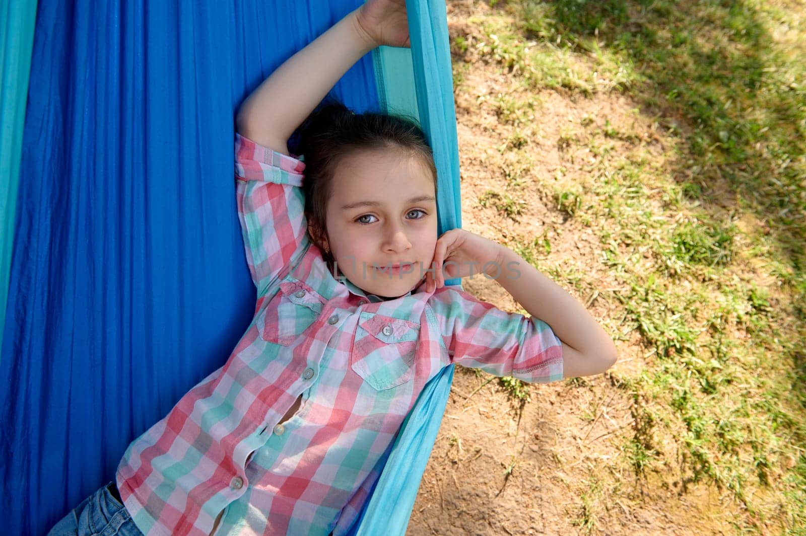 Adorable little child girl smiles looking at camera, relaxing, lying on a blue hammock in the backyard or summer camp, enjoying her weekend outdoors. People. Kids. Leisure activity. Lifestyle.