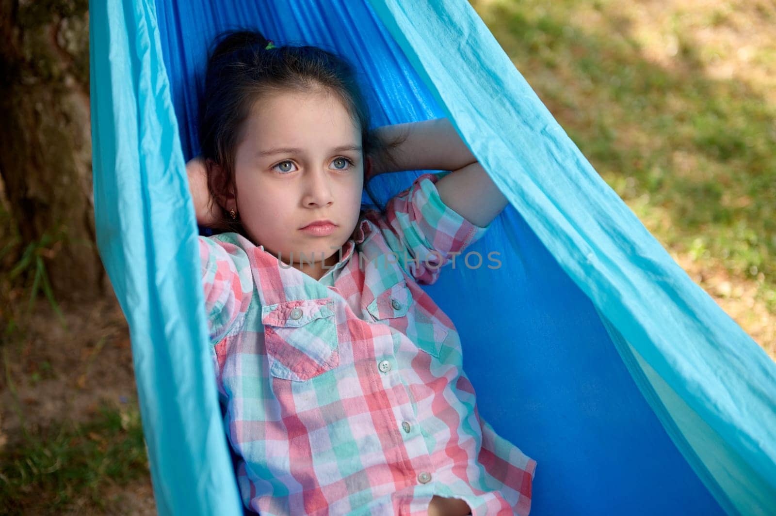Close-up portrait of lovely little child girl dressed in casual denim and pink checkered shirt, relaxing, lying on a blue hammock in the backyard or summer camp, enjoying her weekend outdoors