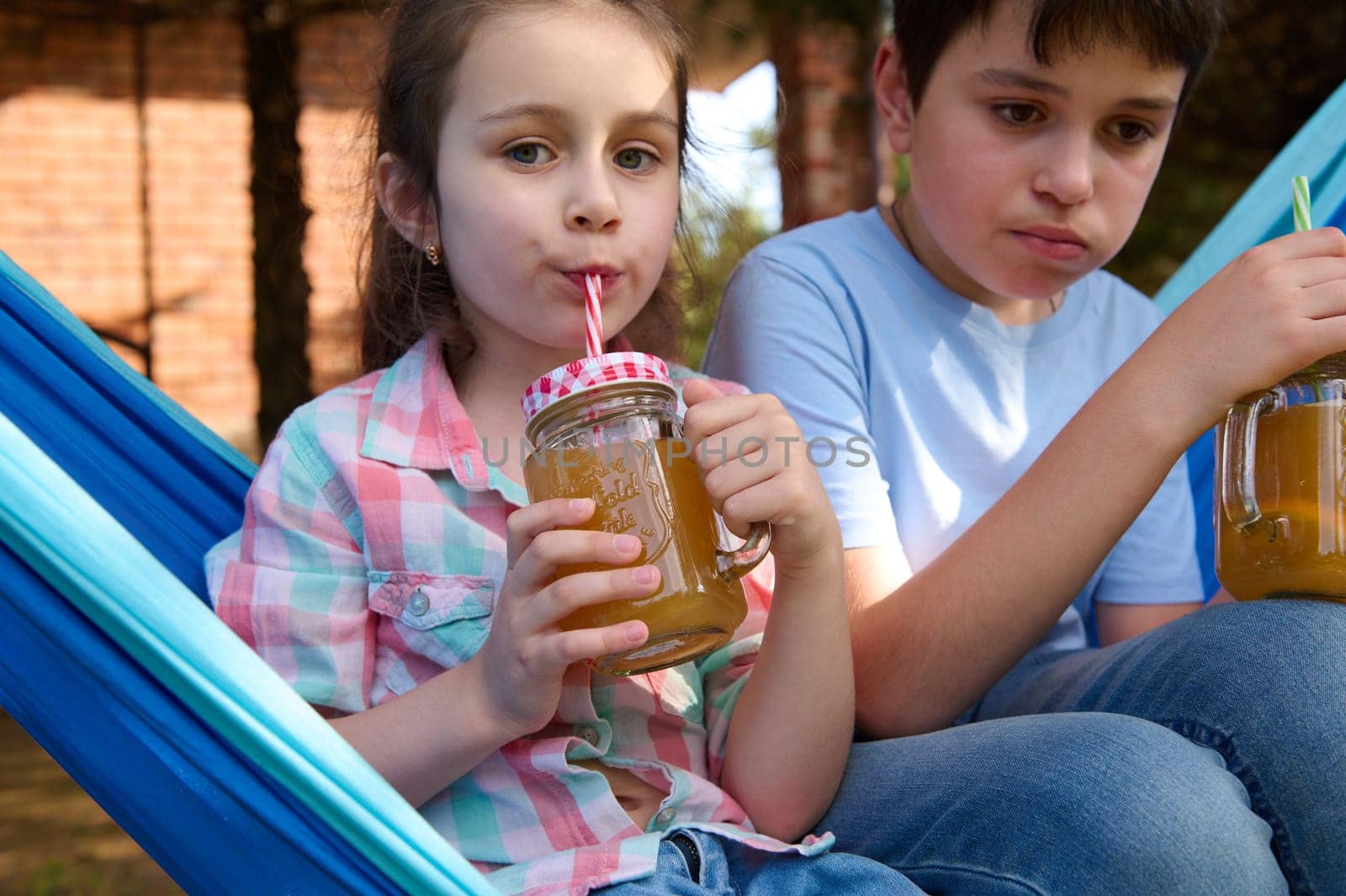 Close-up adorable school kids drinking smoothie while relaxing on hammock in the backyard. Children. Leisure. Lifestyle by artgf