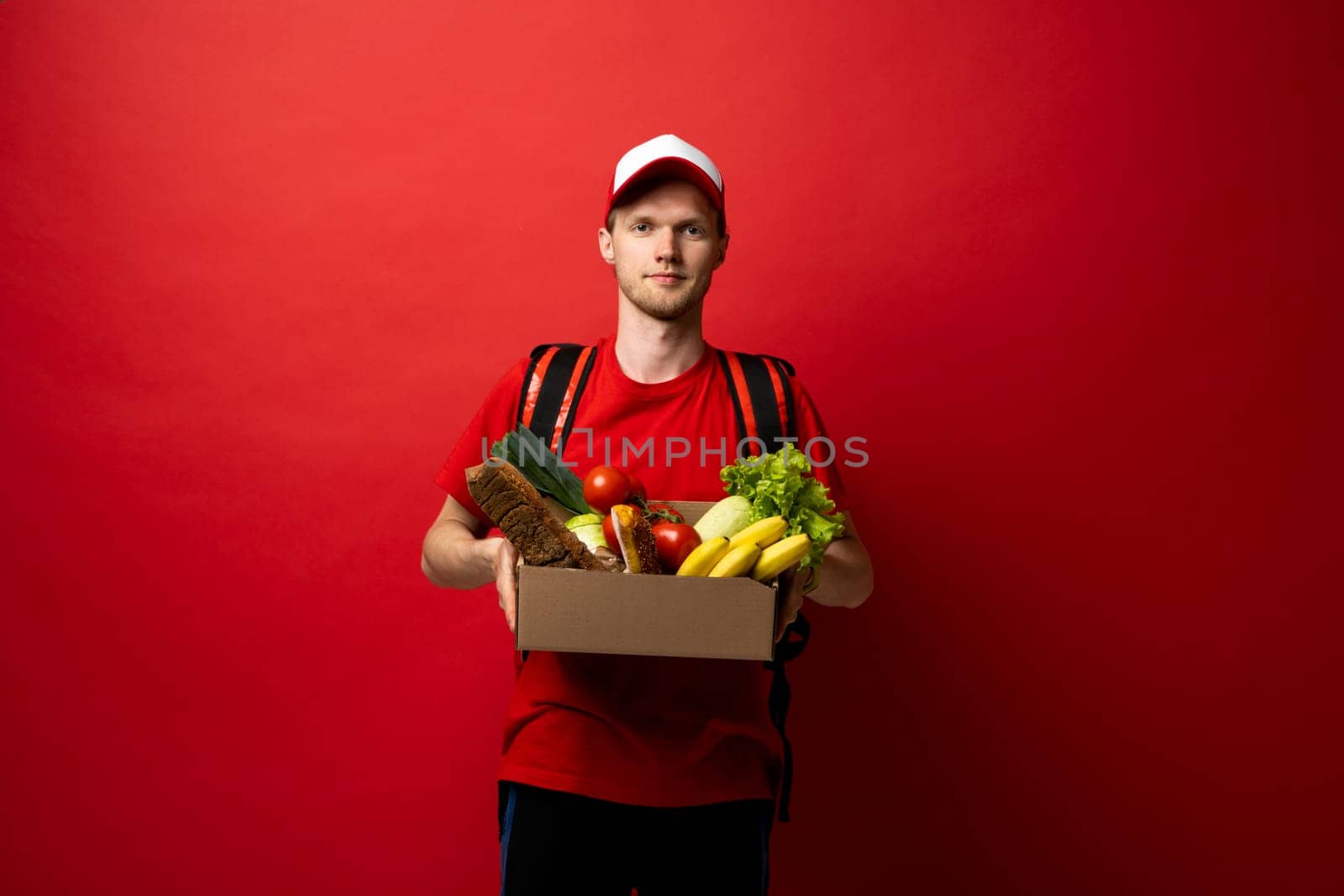 Delivery man holding paper box full of groceries over the white wall. Online supermarket. Food delivery service