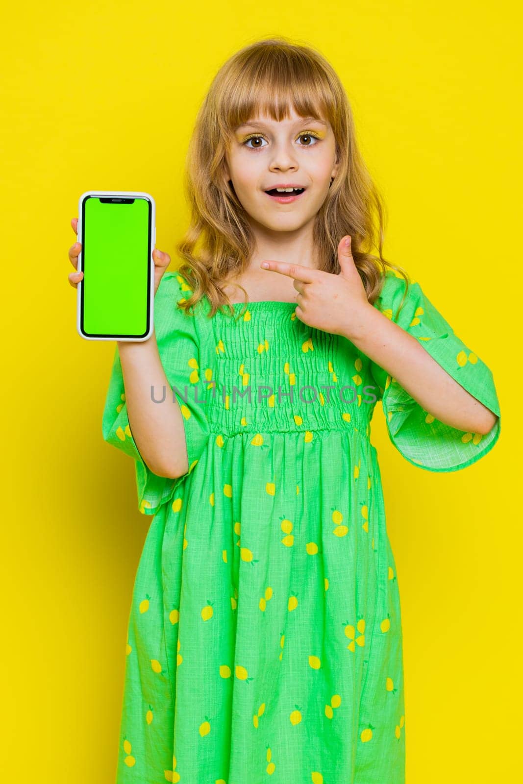 Blonde child girl kid hold smartphone with green screen chroma key mock up recommend good game play application promotional sale offer. Little preteen children isolated on yellow background. Vertical