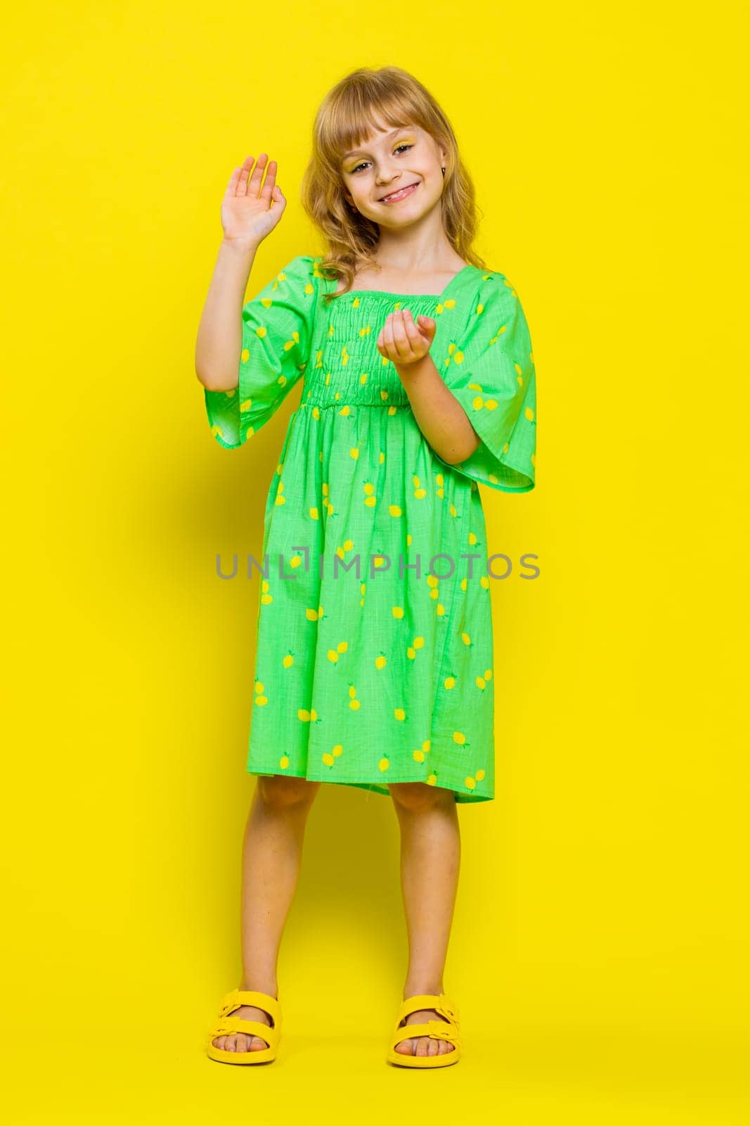 Young blonde school girl smiling friendly at camera and waving hands gesturing hello or goodbye, welcoming with hospitable expression. Preteen female child kid isolated on yellow background. Vertical