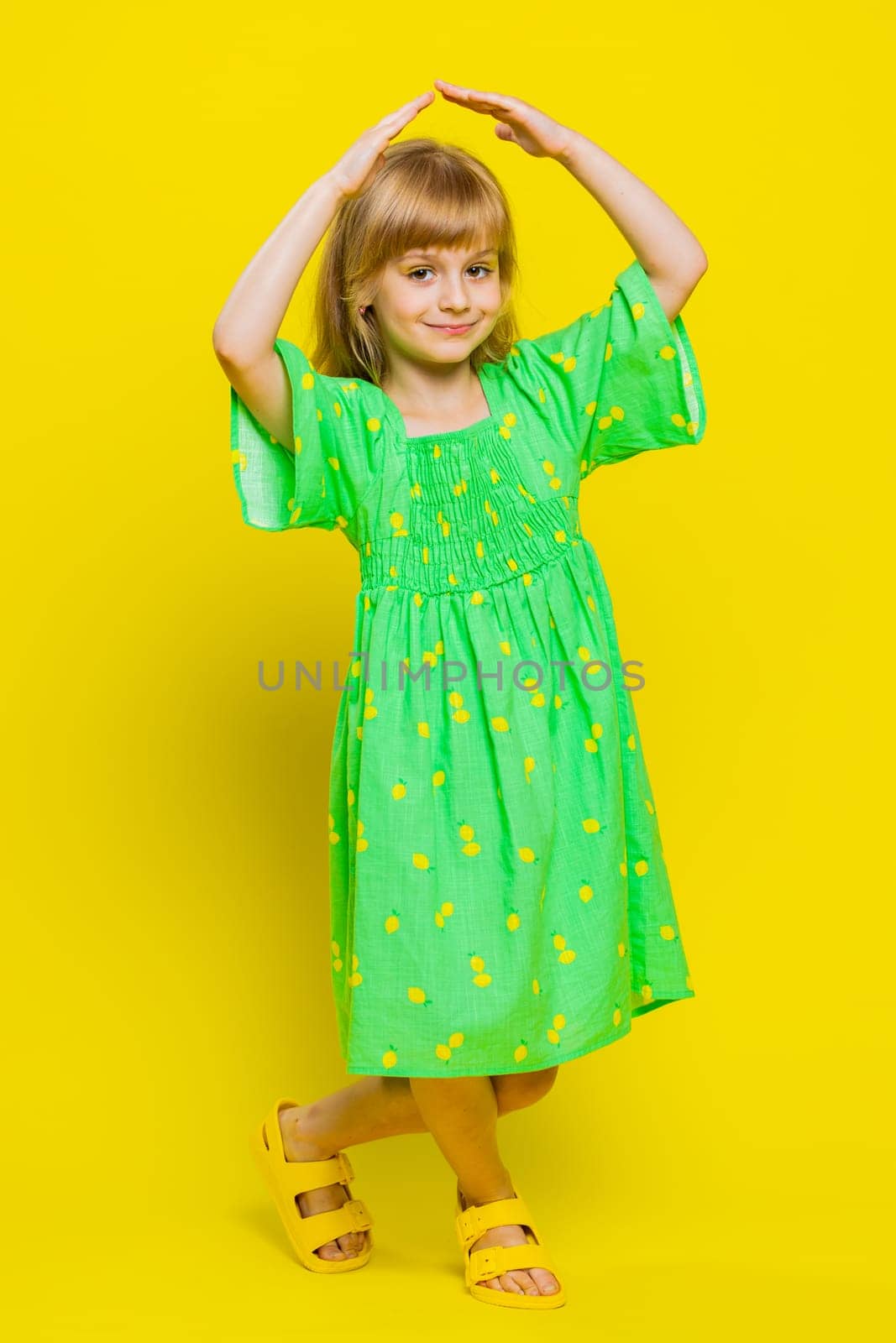 Young blonde child girl kid playing childish catching up game, feeling in safe making roof above head with hands, insurance, security service. Little preteen children on yellow background. Vertical