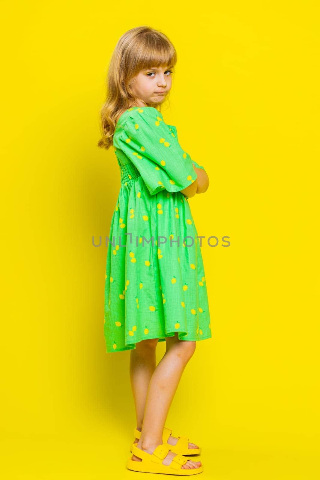 Cheerful funny young blonde school girl making faces at camera, fooling around, joking, aping with silly face, teasing, bullying, abuse. Preteen child kid on yellow background. Vertical