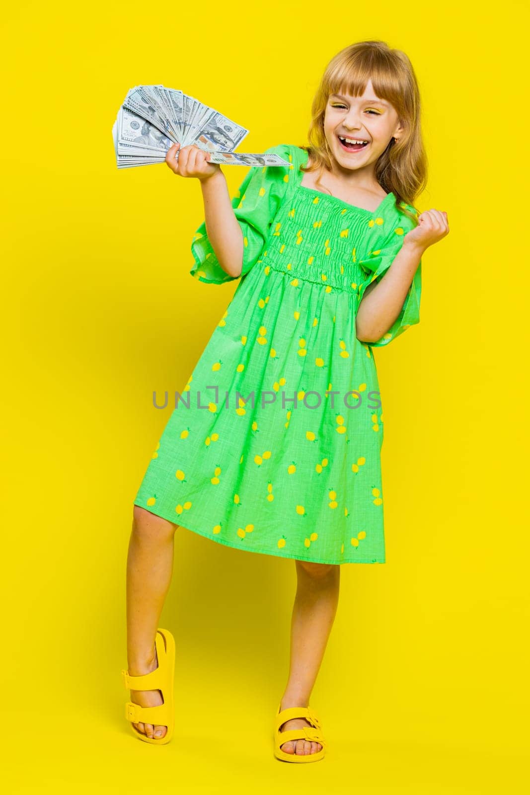 Rich pleased young cute school girl waving money dollar cash banknotes bills like a fan, success, lottery winner, big income, wealth. Teen blonde female child kid on studio yellow background. Vertical