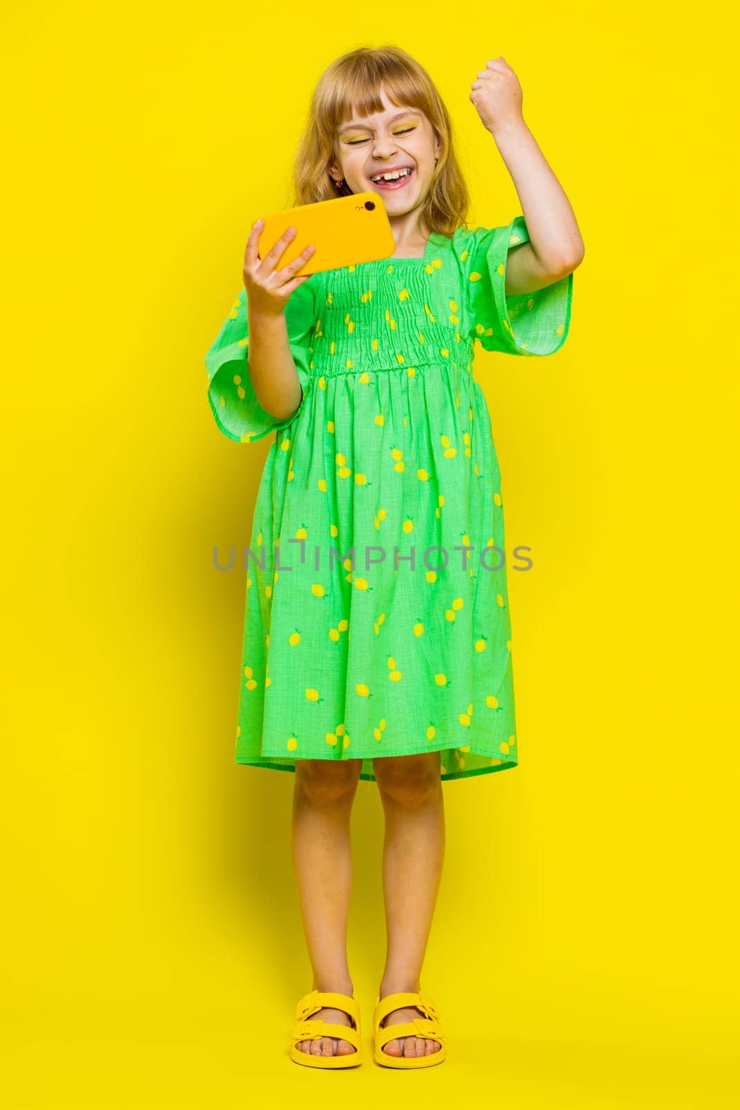 Worried funny young school girl enthusiastically playing racing or shooter video games on smartphone. Preteen child kid using smartphone gadget app with drive simulator on yellow background. Vertical