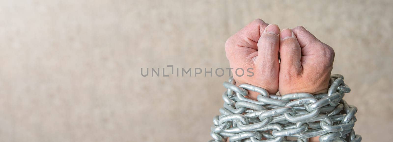 The rough hands of a worker, a white male are wrapped in a chain as a concept of slavery or imprisonment. Close-up photo of the hands of a slave with a chain on a gray background with copy space