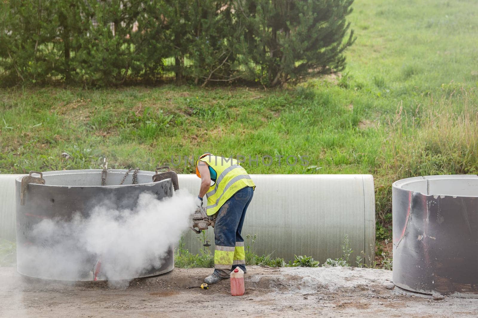 Installing a concrete pipe in the ground to collect rainwater. The worker cuts holes in the concrete part of the pipe to connect to the storm drain. A man in construction clothes is sawing concrete. by SERSOL