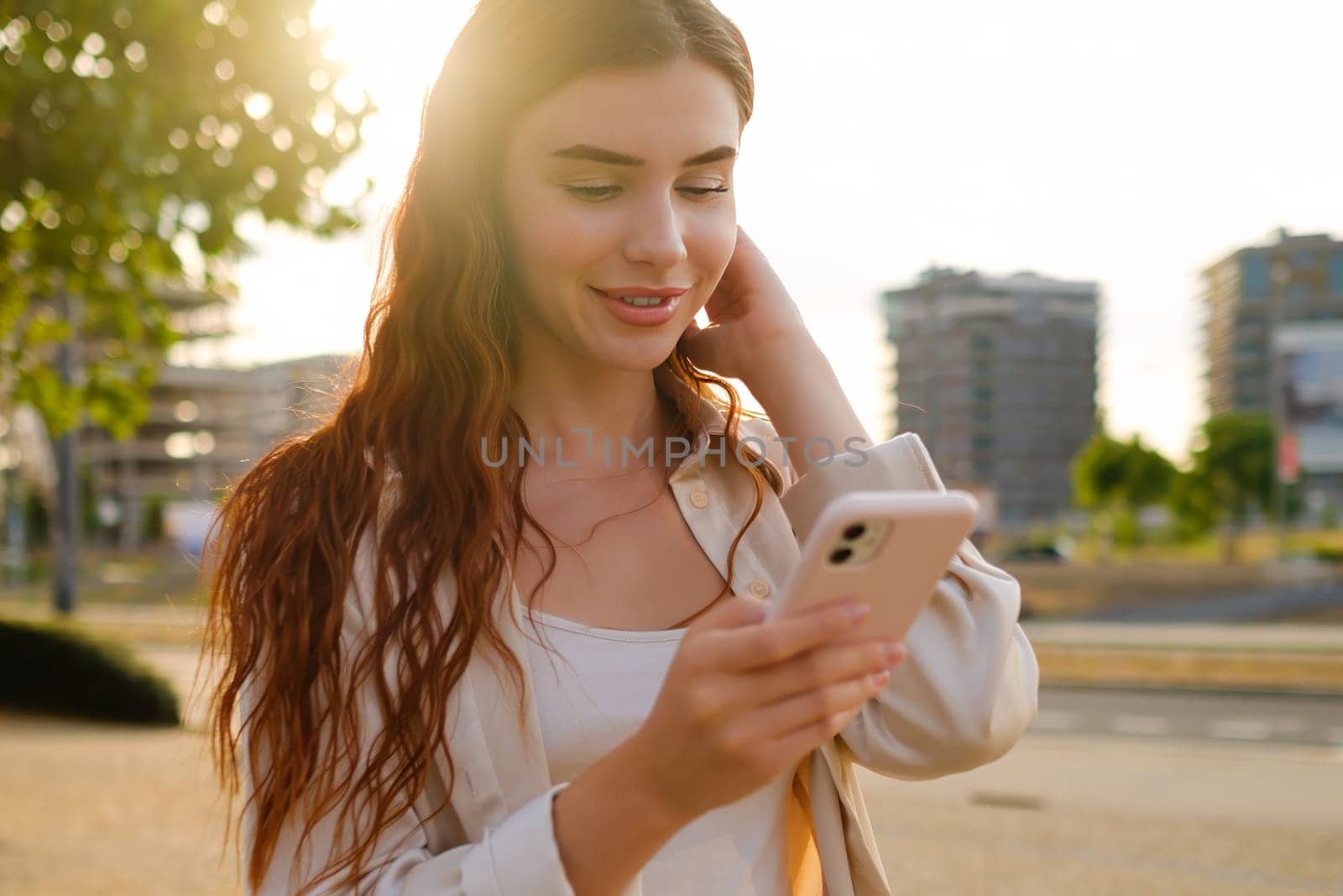 Romantic pretty young red haired woman standing on the street and texting using her smartphone at sunset