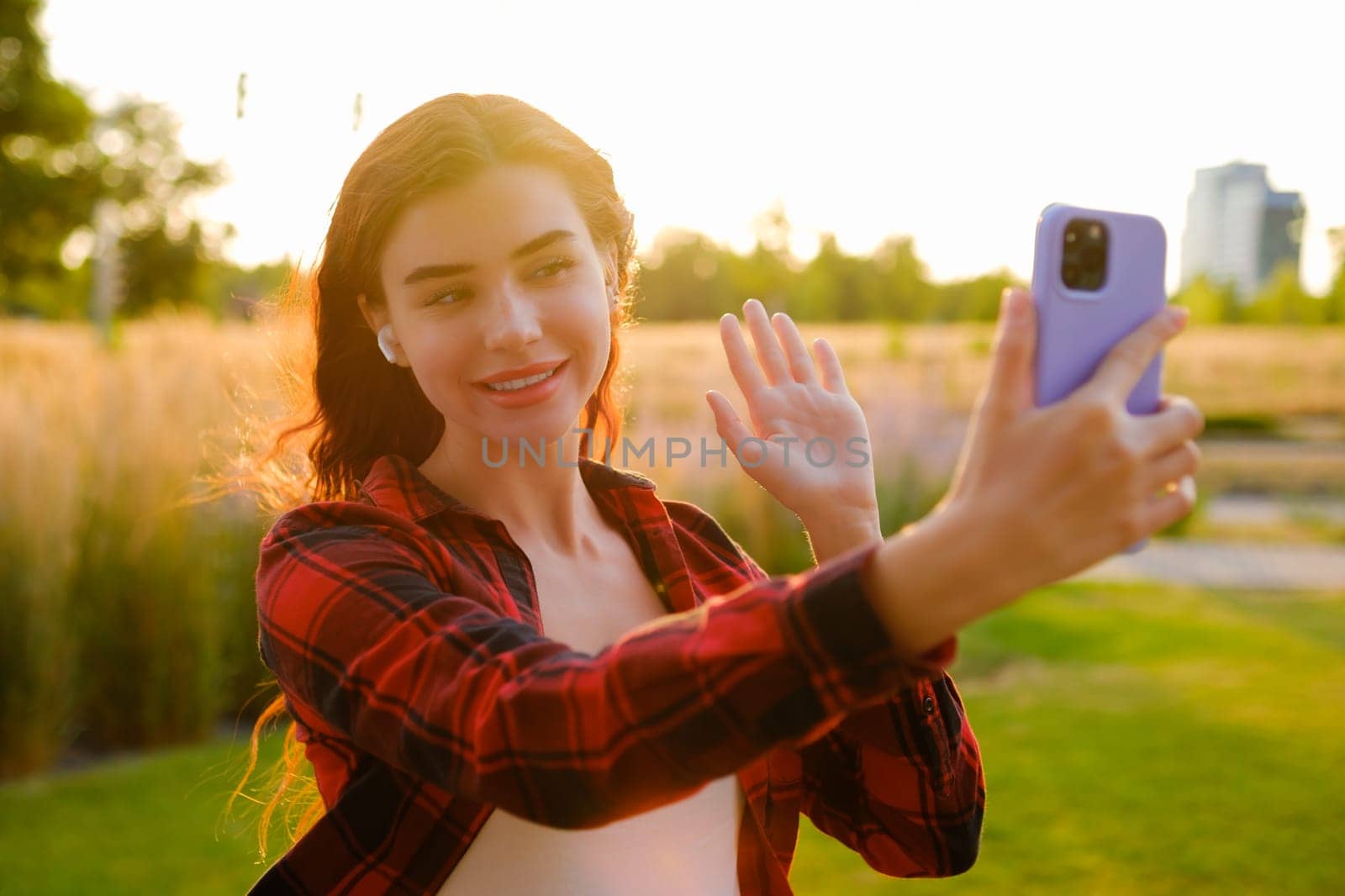 A portrait of a happy red-haired young blogger in a plaid shirt warmly greets her followers, recording a captivating story using her mobile phone at sunset