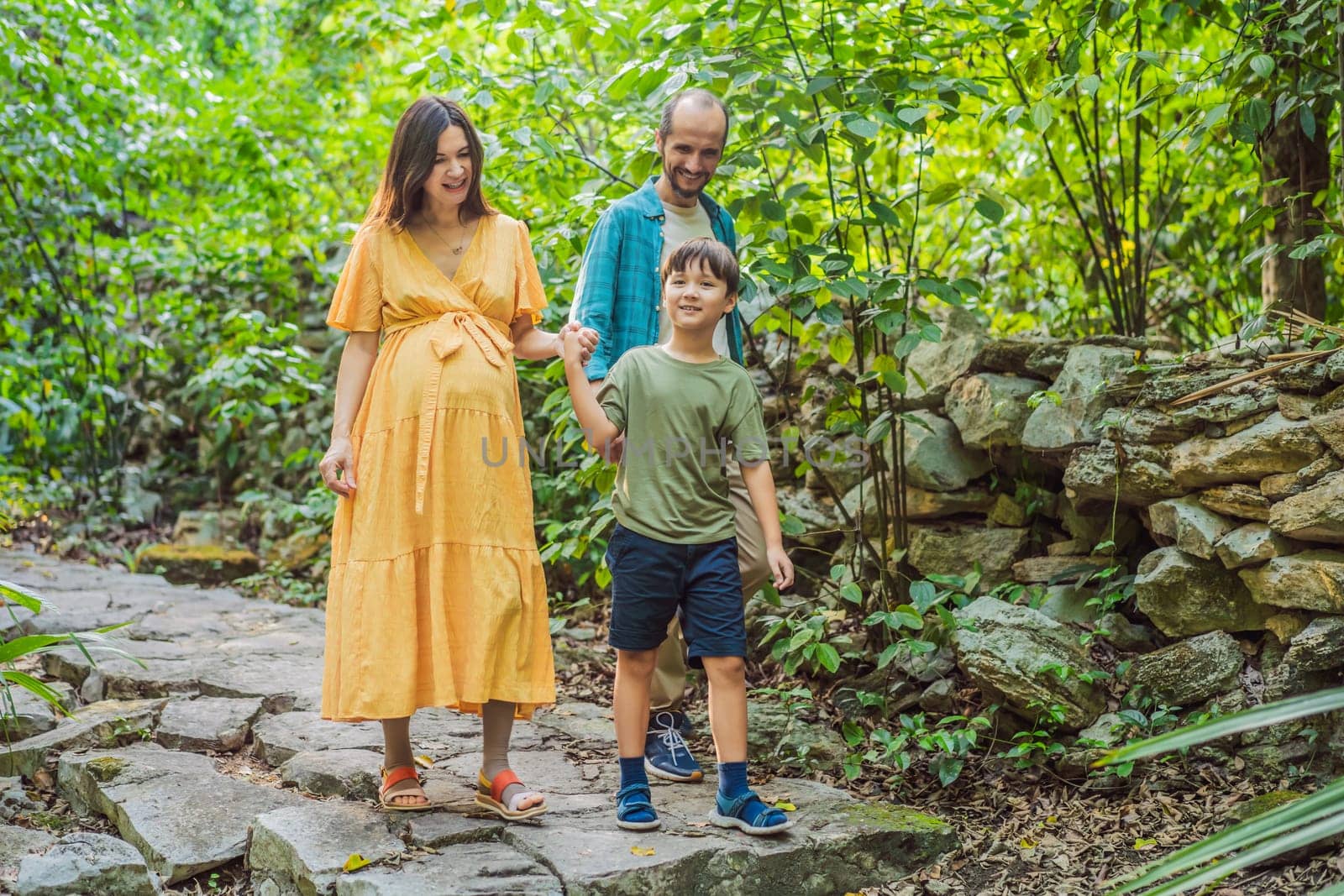 A loving family enjoying a leisurely walk in the park - a radiant pregnant woman after 40, embraced by her husband, and accompanied by their adult teenage son, savoring precious moments together amidst nature's beauty. Pregnancy after 40 concept.