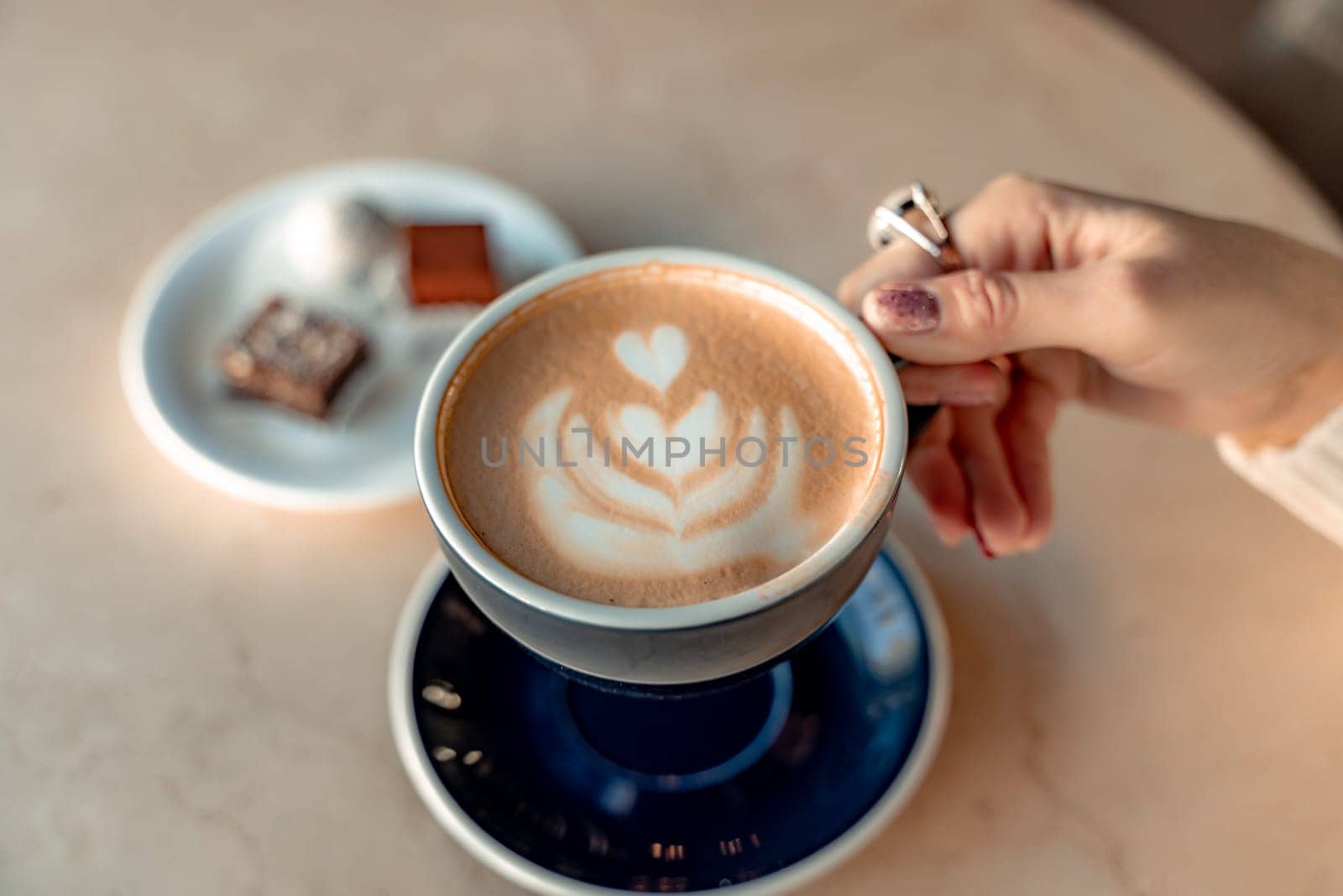 A cup of fresh cappuccino coffee in the hands of a woman on a fashionable background of a white marble table, next to a plate with sweets. Coffee addiction. Top view, flat lay