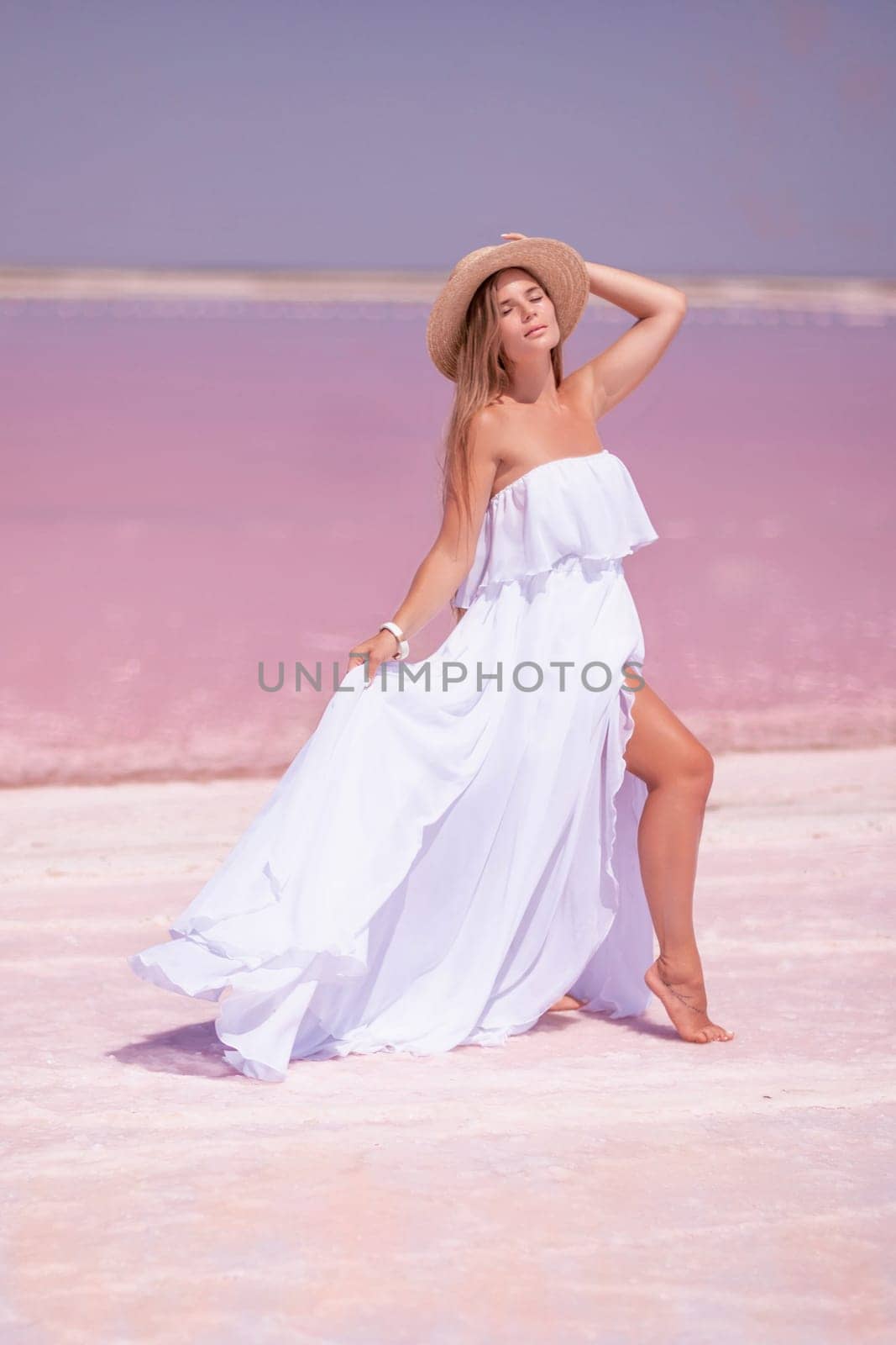 Woman in pink salt lake. She in a white dress and hat enjoys the scenic view of a pink salt lake as she walks along the white, salty shore, creating a lasting memory