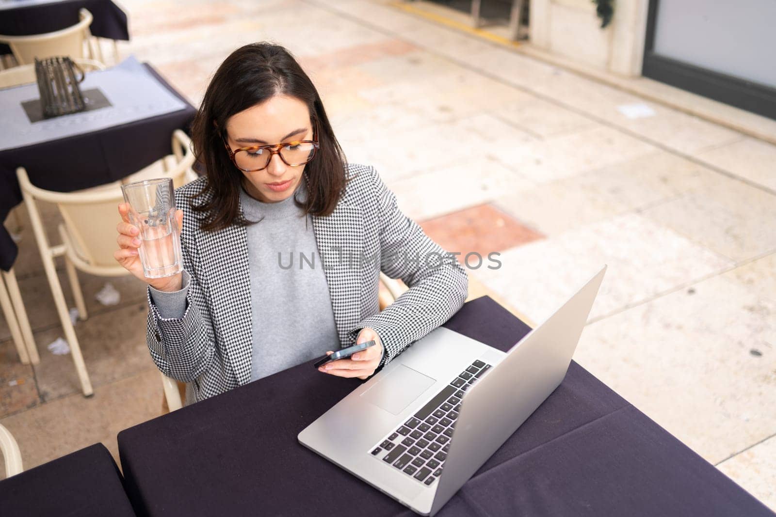 Freelancer using smartphone and laptop sitting outdoor cafe drink water wearing glasses, working in cafe on laptop, holding mobile phone in hands, drinking water from glass. High angle view