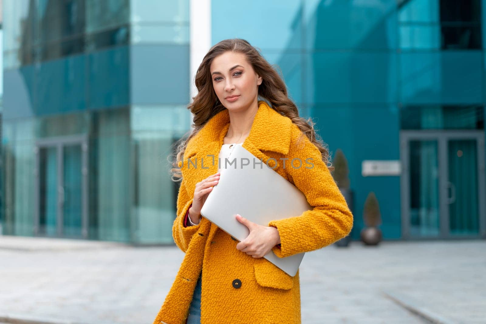 Business woman with laptop dressed yellow coat standing outdoors corporative building background Caucasian female business person on city street