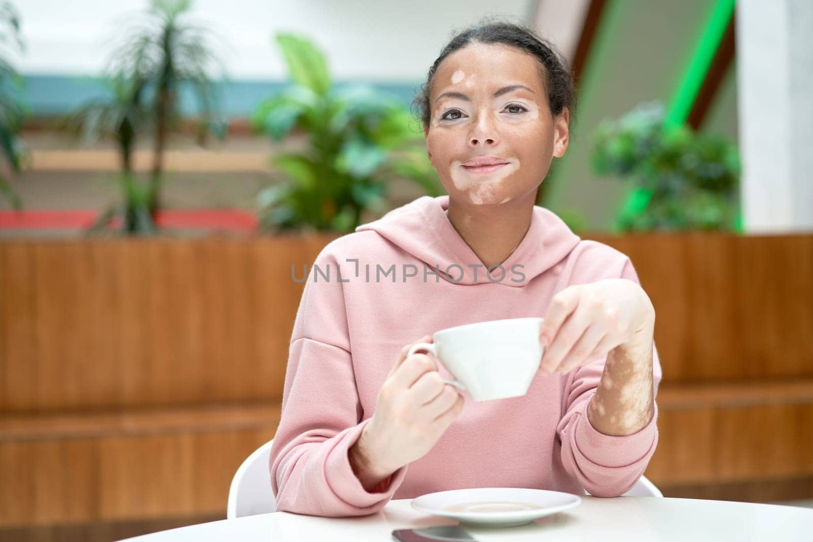 Black african american woman with vitiligo pigmentation skin problem indoor dressed pink hoodie drink tea white mug