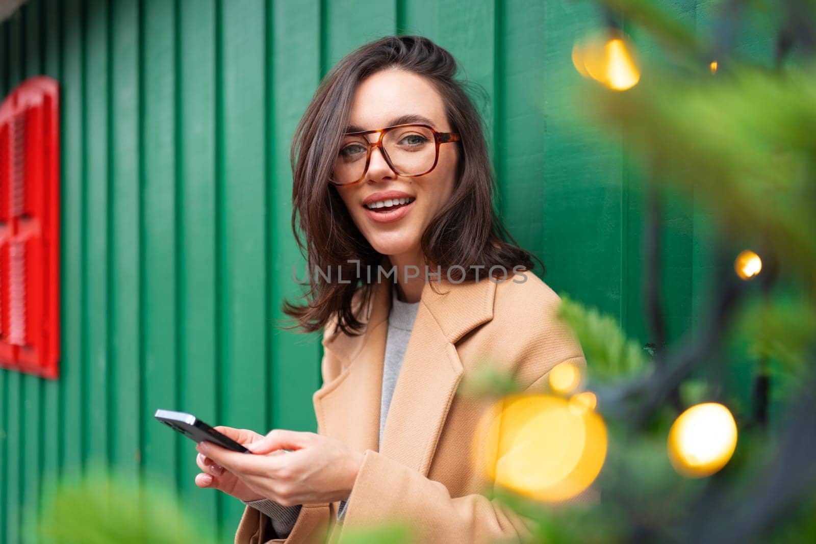 Close up smiling woman using smartphone, standing near Christmas tree, chatting or shopping online, choosing gifts, happy female spending winter holidays with mobile device outside dressed trench coat