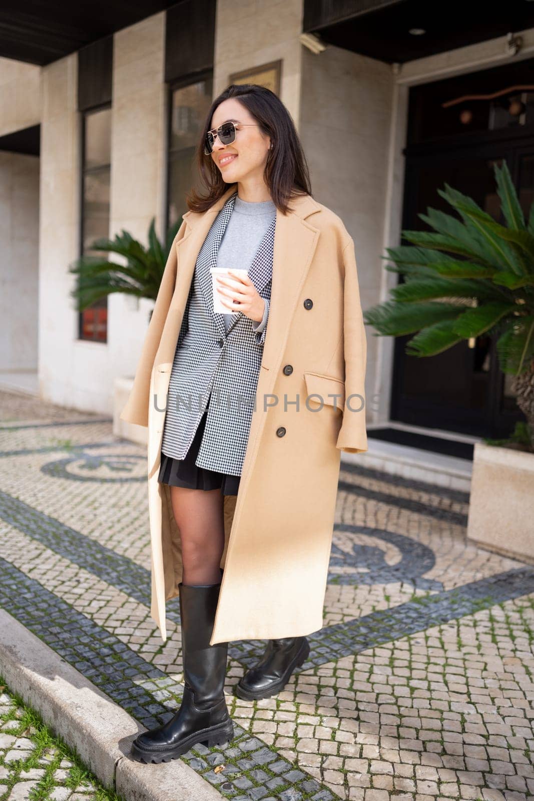 Happy caucasian girl walking with cup of coffee in city on sunny day. Brunette woman wears stylish jacket coat and sunglasses come out from building entrance. Side view vertical photo