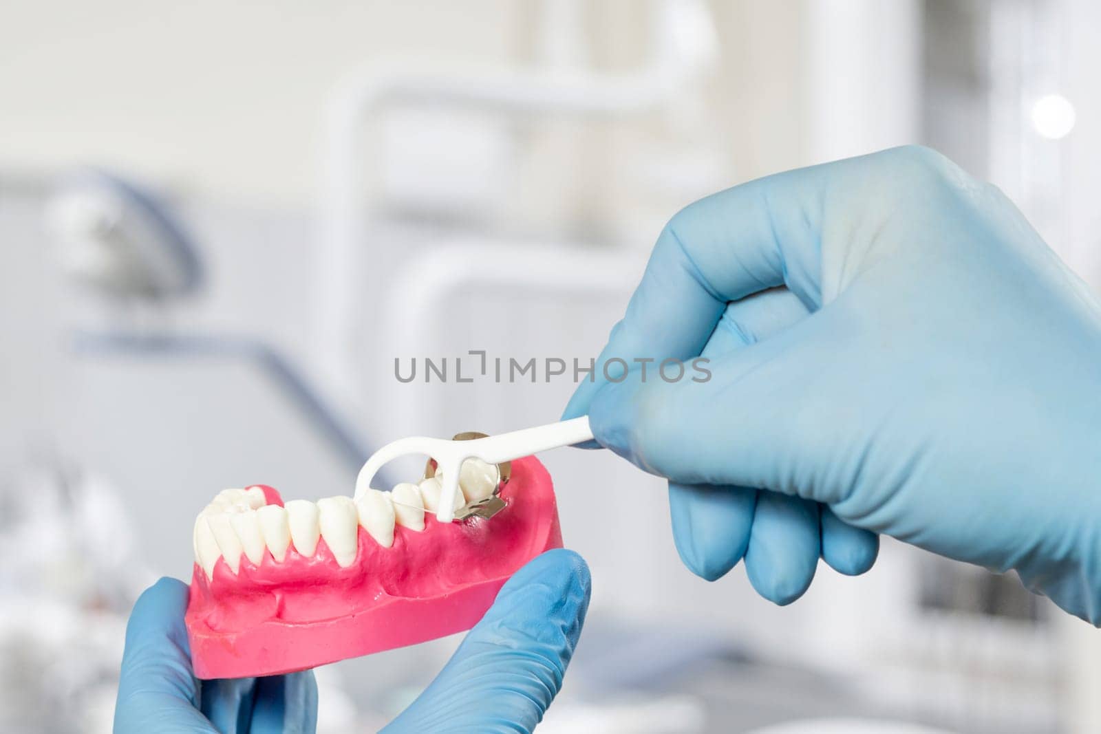 Close-up view of dentist's hands with a human jaw layout and a floss toothpick. Dental office on the background.