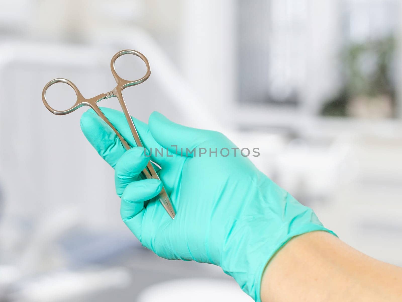 Woman's hand in a latex glove with a stainless steel needle holder. Dental office on the background.
