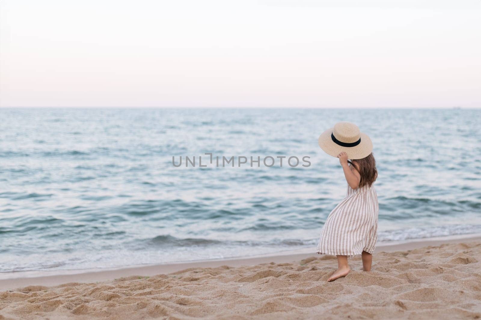 Little girl in a straw hat on the beach by the sea, back view.