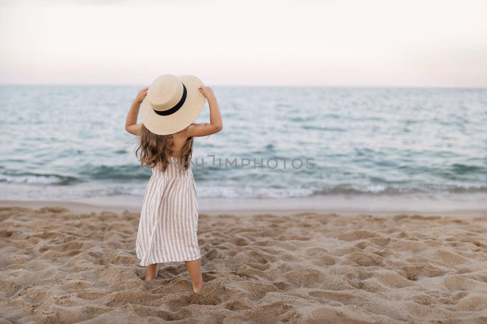 Little girl in a straw hat on the beach by the sea, back view.