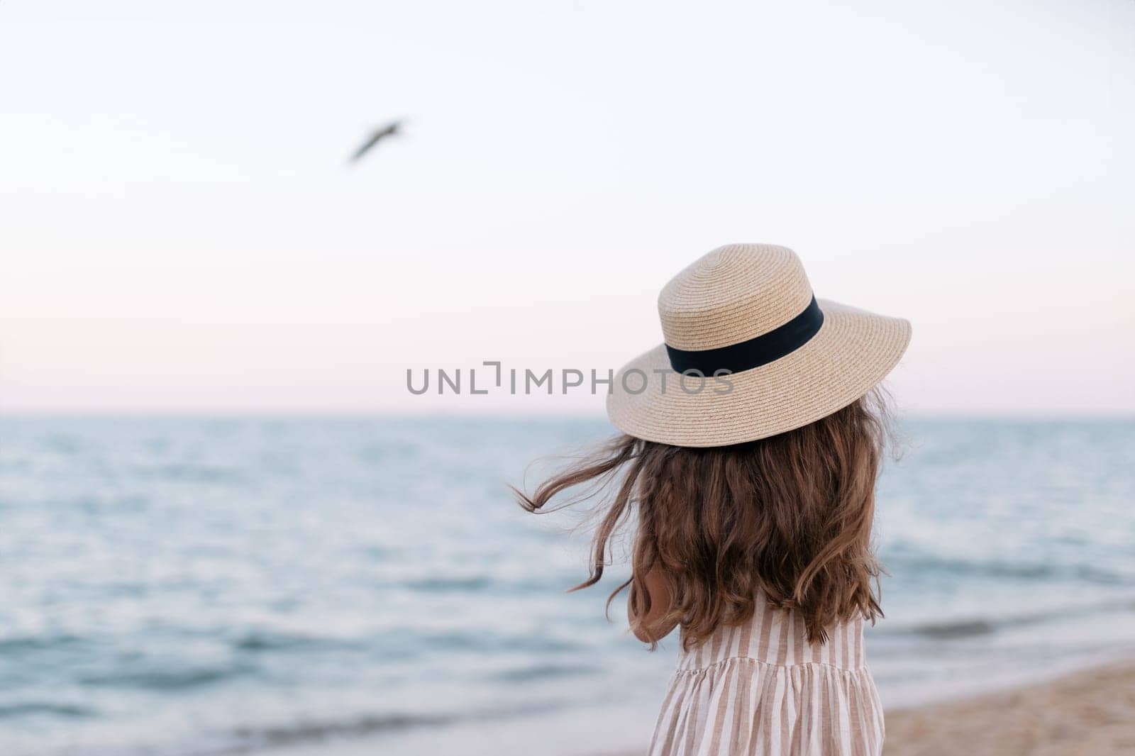 Close-up of a girl in a straw hat looks at the sea into the distance on a sunny day. The sea wind is blowing. Background of sea waves, seagulls. Pleasant little girl enjoying seascape view.