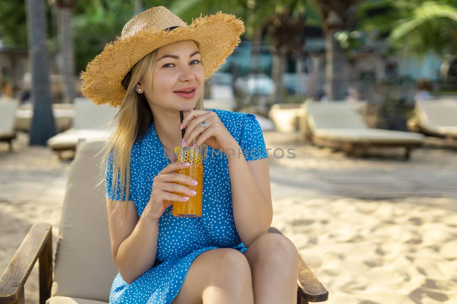 Fashion young woman drinking cocktail in beach bar. Cheerful attractive blonde woman in a dress smiles and drinks a cocktail outdoors. Happy woman enjoying summer time at the beach.
