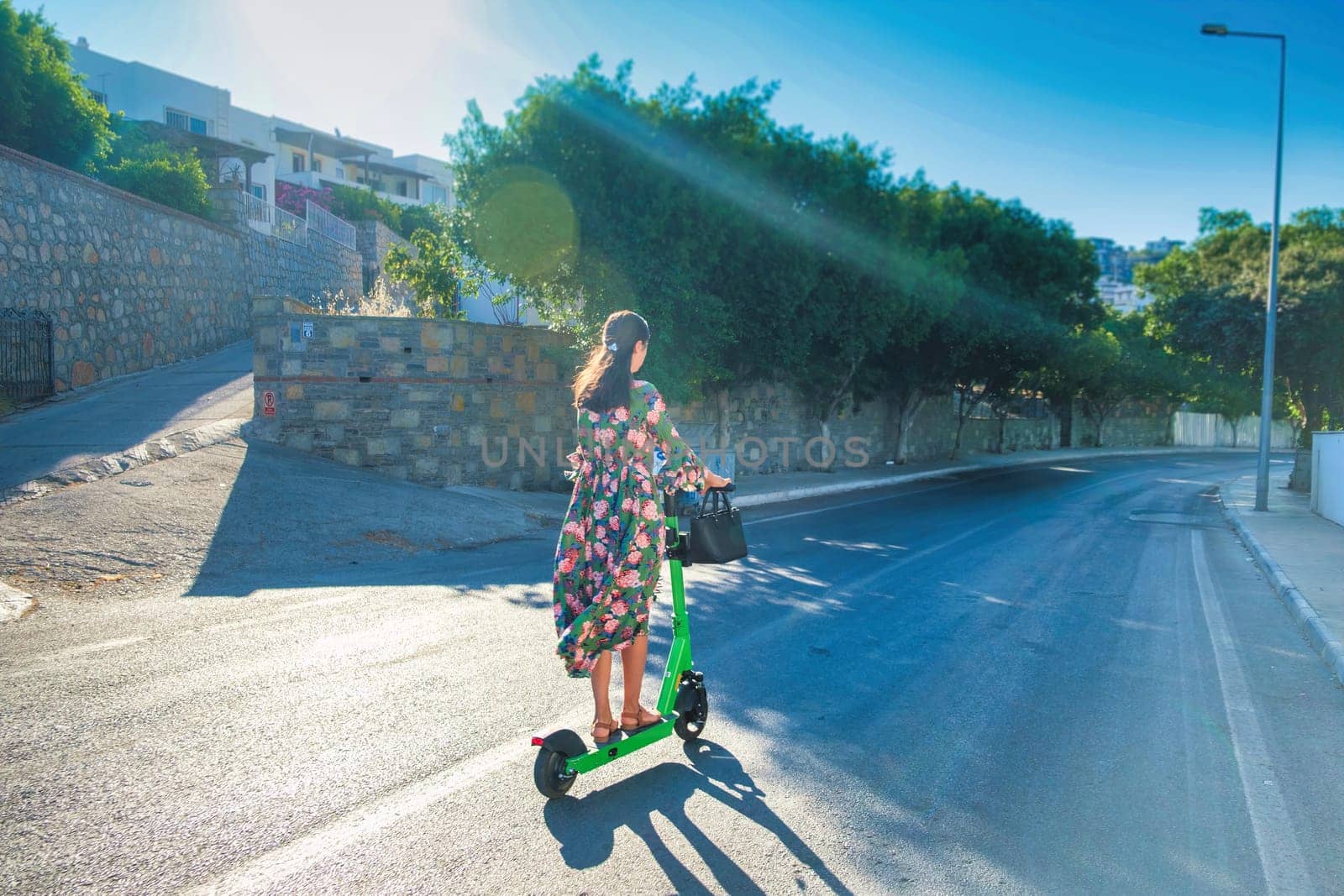 Young beautiful woman in dress rides an electric scooter on empty asphalt road. Electric urban transportation concept image by igor010