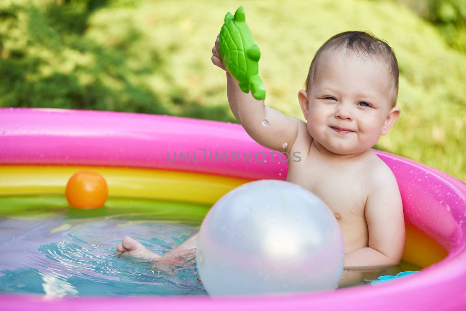 Happy cute baby girl playing with ball in kid inflatable pool in summer day in the backyard