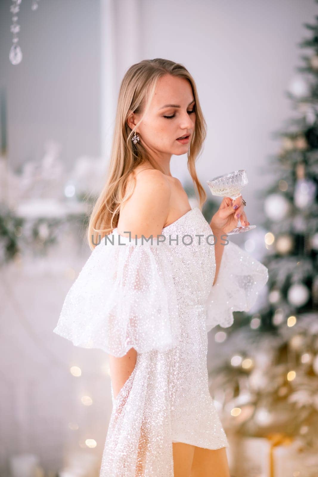 The blonde in the Christmas room. A beautiful blonde woman in a shiny light short dress with a train stands in a beautiful bright room decorated with a festive interior with a Christmas tree