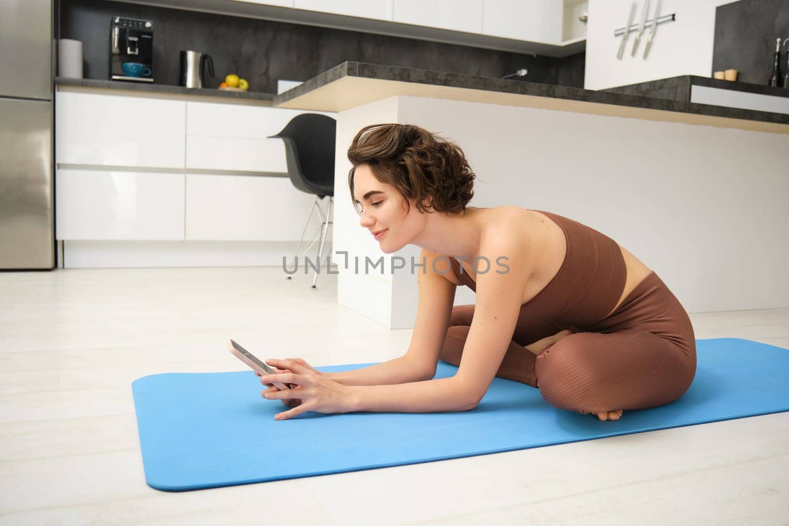 Portrait of fitness woman in leggings and sportsbra, sits in kitchen and does workout on yoga mat, looks at mobile phone, exercise from home with smartphone app.