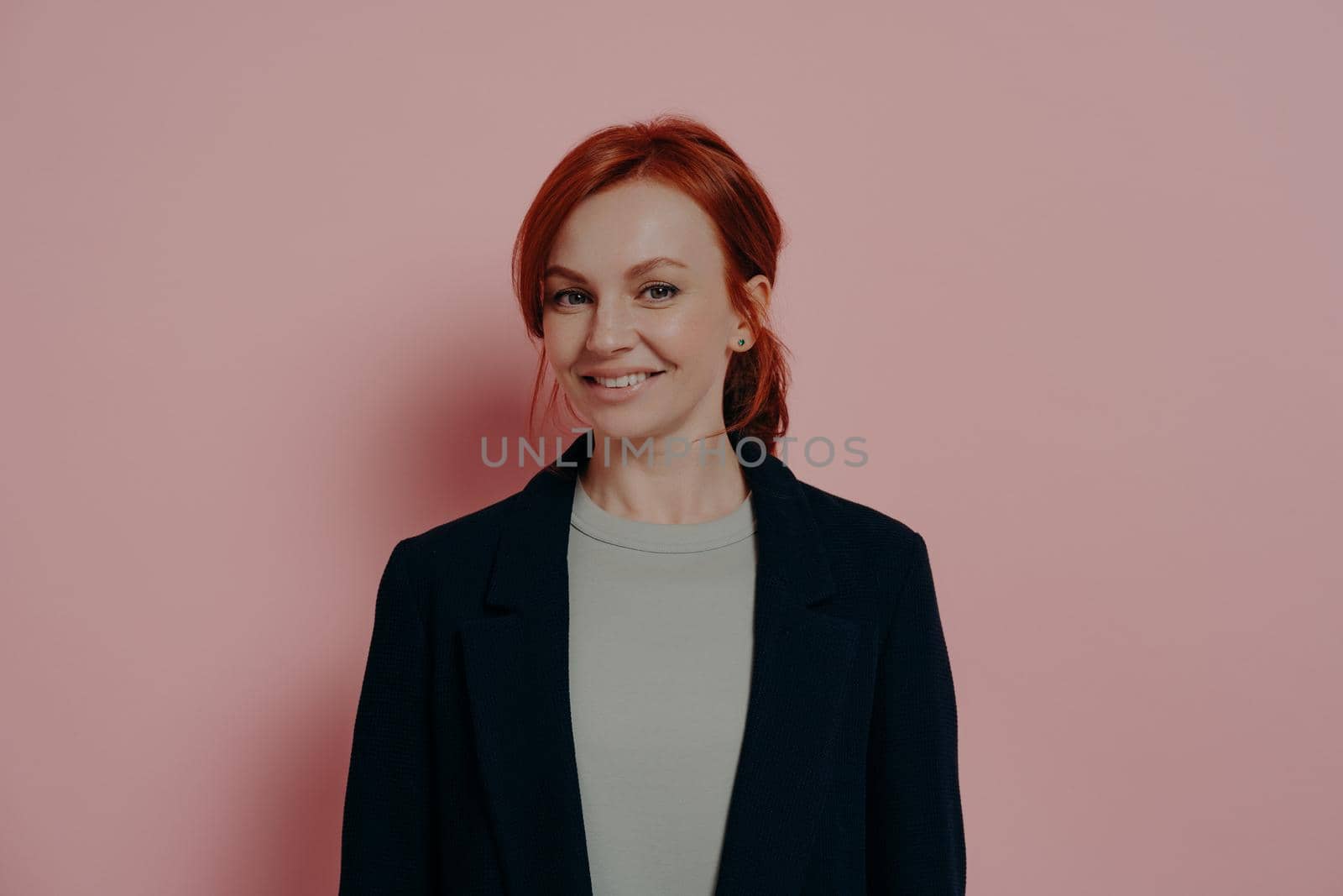 Successful pretty red-haired woman looking at camera with confidence and pleasant smile, standing isolated on rosy background in studio, stylish ginger female model in casual outfit smiling cheerfully