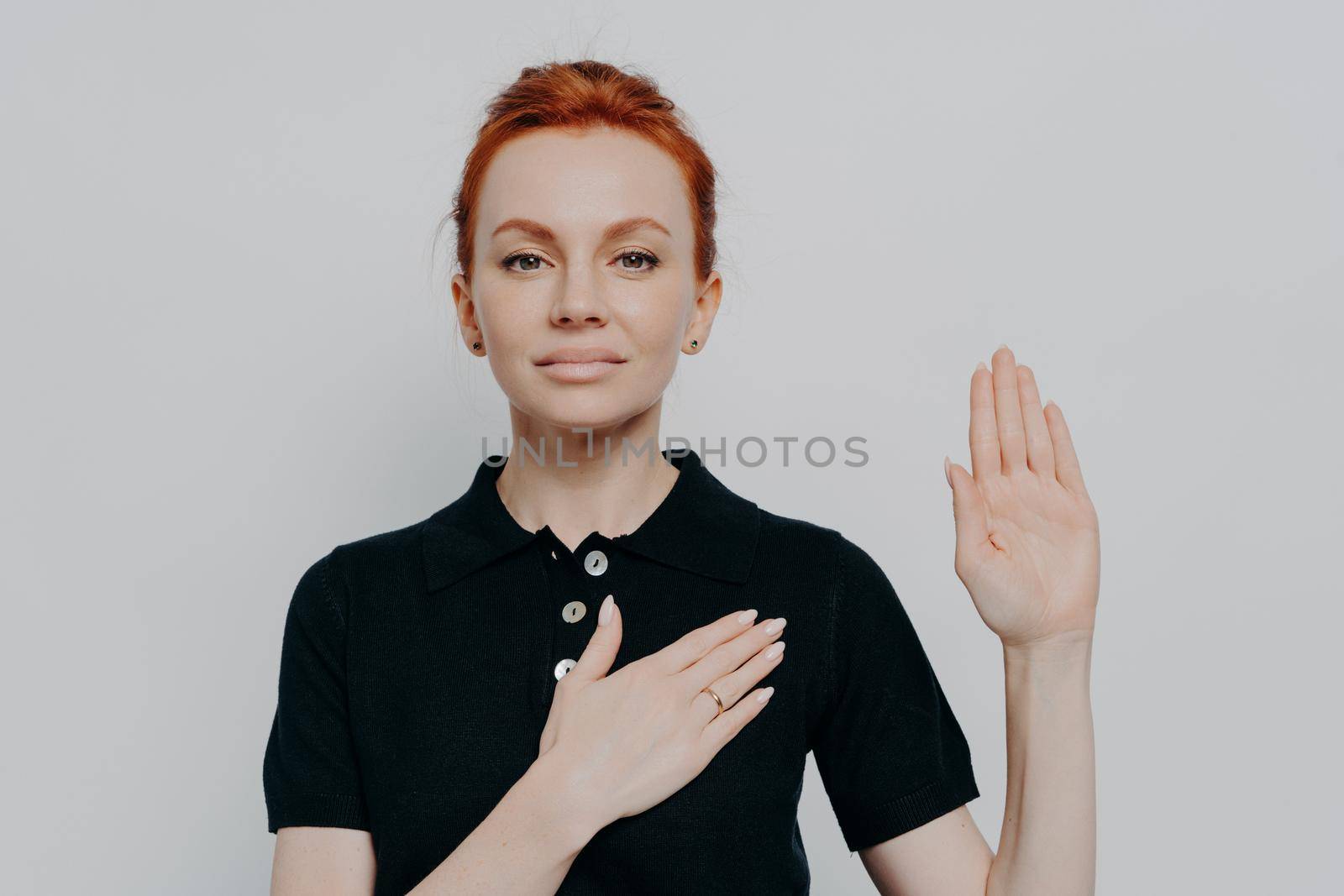 Beautiful red-haired patriotic woman wearing black t-shirt swearing with hand on chest and open palm, making loyalty promise oath while posing isolated over grey studio background