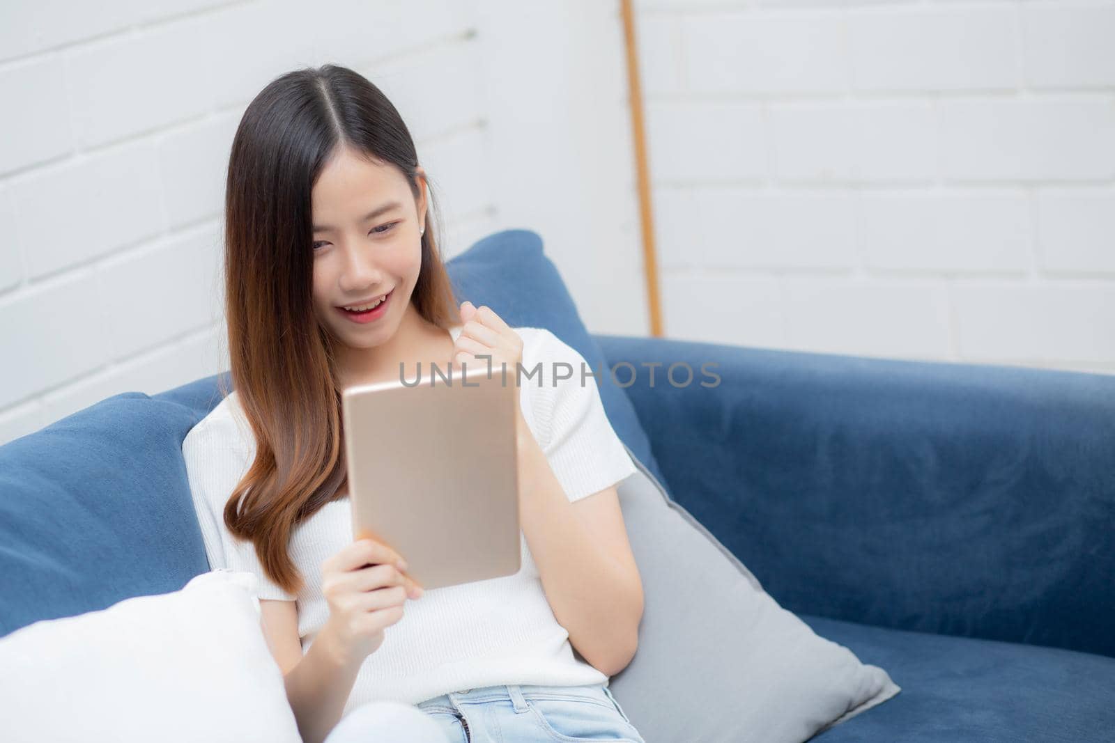 Young attractive asian woman resting using browsing tablet computer on sofa at home, happy girl sitting on couch relax reading digital gadget at house, communication and lifestyle concept.