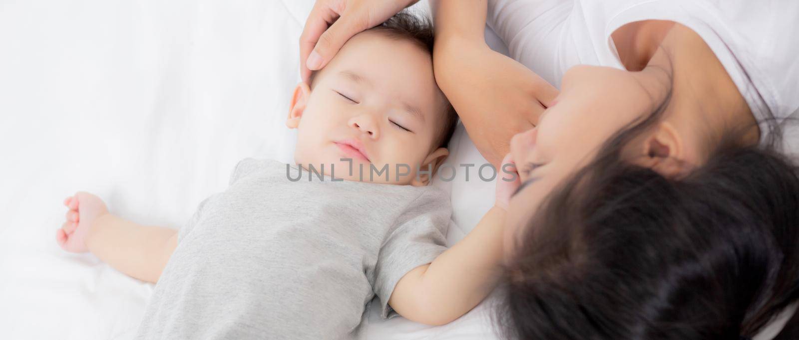 Young asian mother touch head of little baby girl with tender on bed in the bedroom, mom love newborn and care, woman with expression with child together, parent and daughter, family concept.