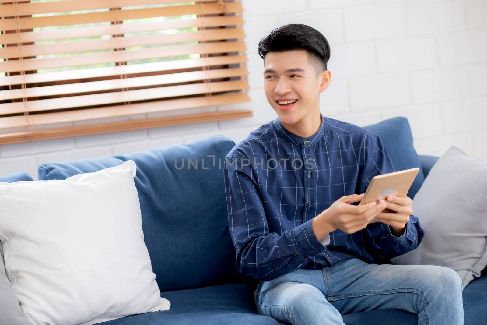 Young attractive asian man resting using browsing tablet computer on sofa at home, happy male sitting on couch relax reading digital gadget at house, communication and lifestyle concept.