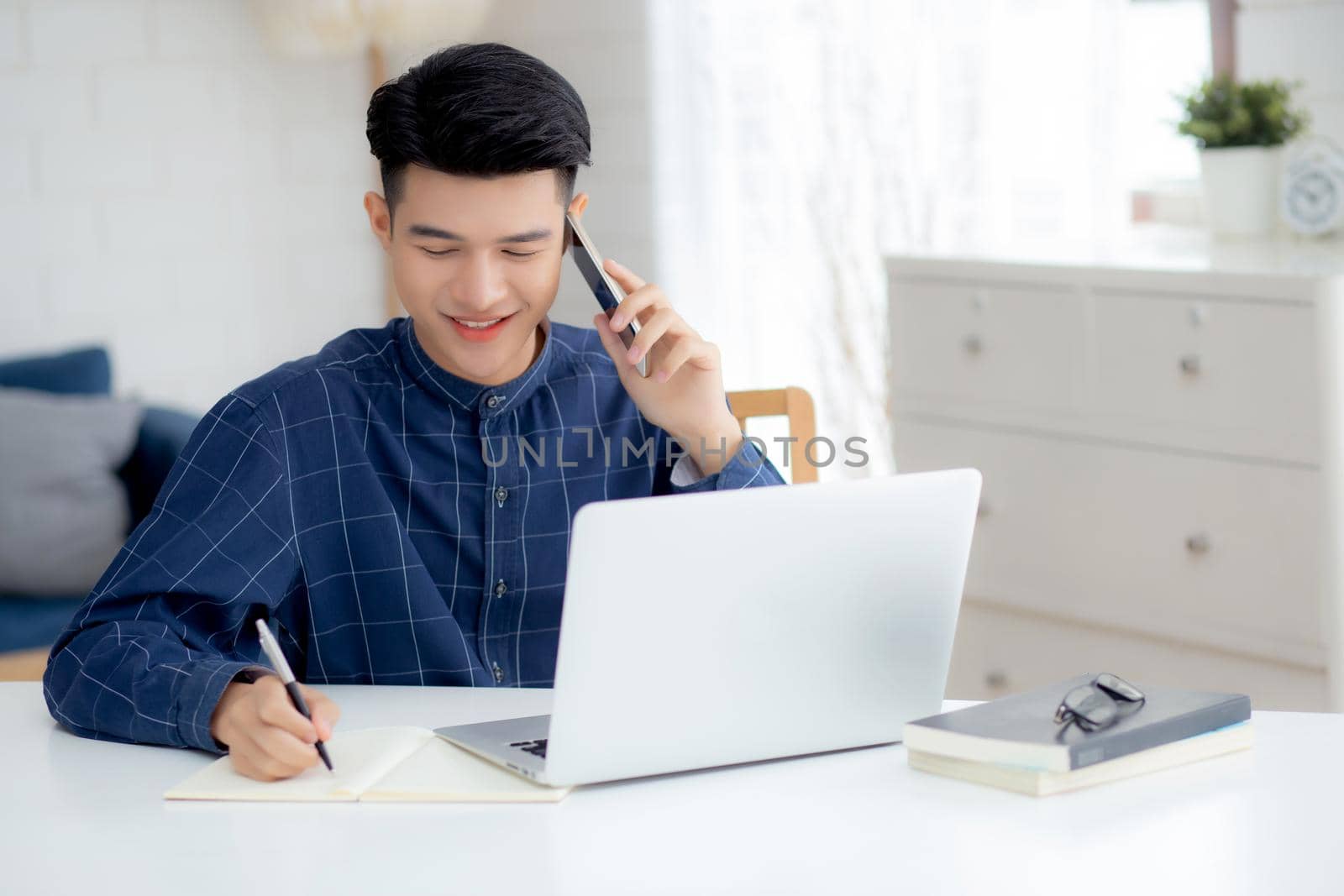 Young asian man talking phone and work from home with laptop computer, freelance male writing on notebook and speak on smartphone, stay home, domestic life, business and communication concept. by nnudoo