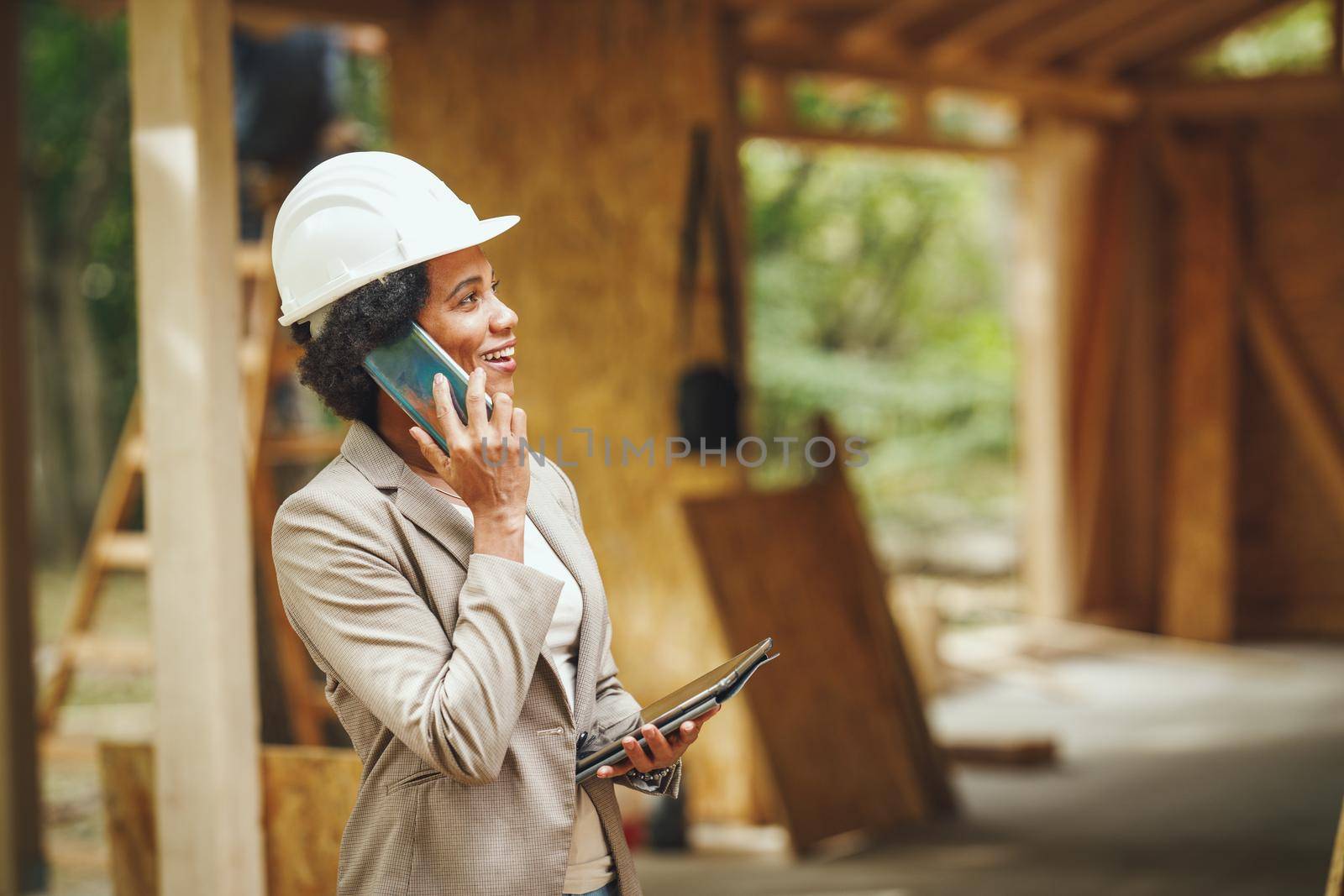 Shot of an African female architect with white helmet using a smartphone and checking construction site of a new wooden house.