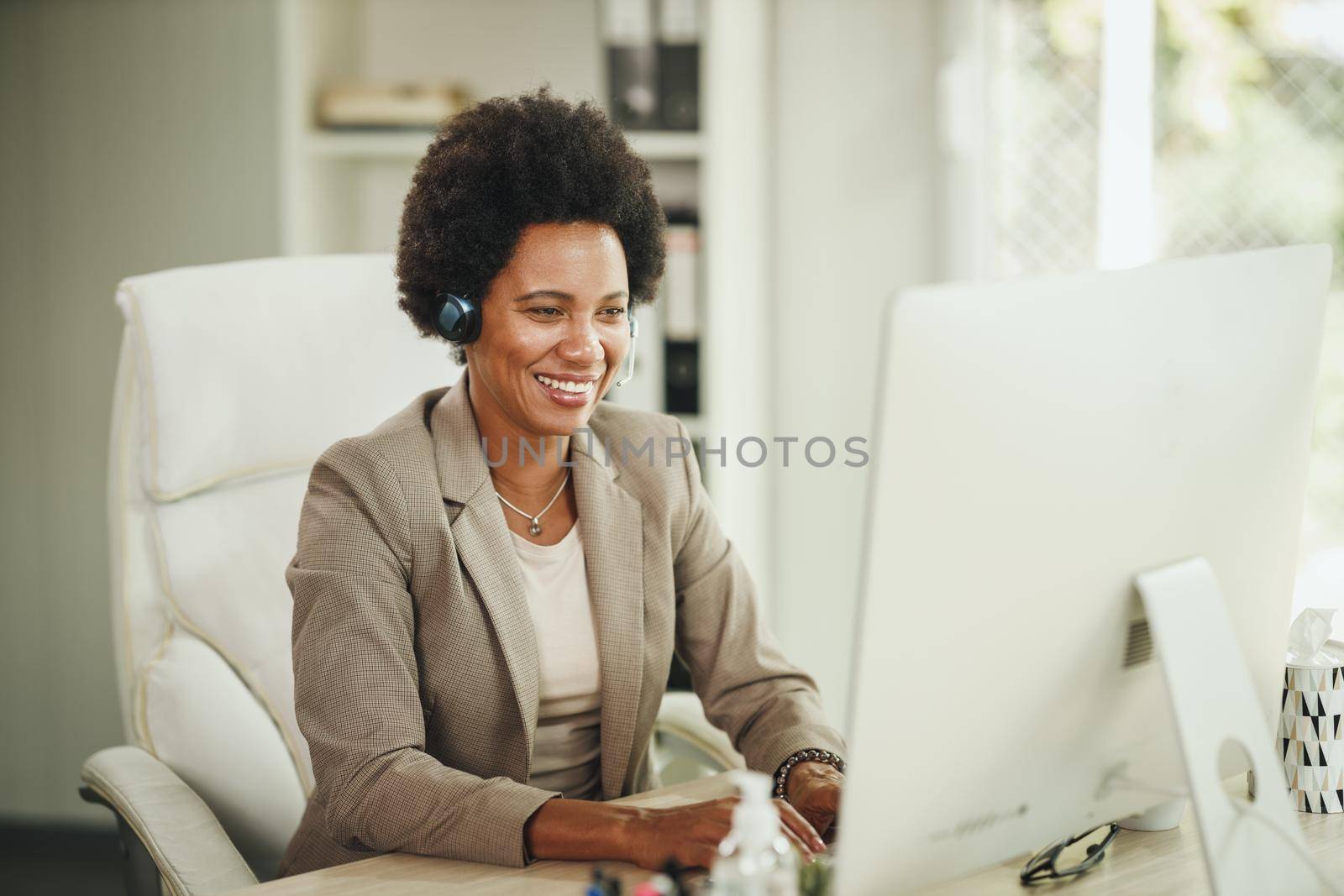 Shot of an attractive African businesswoman sitting alone in her home office with headphones and working on computer during COVID-19 pandemic.