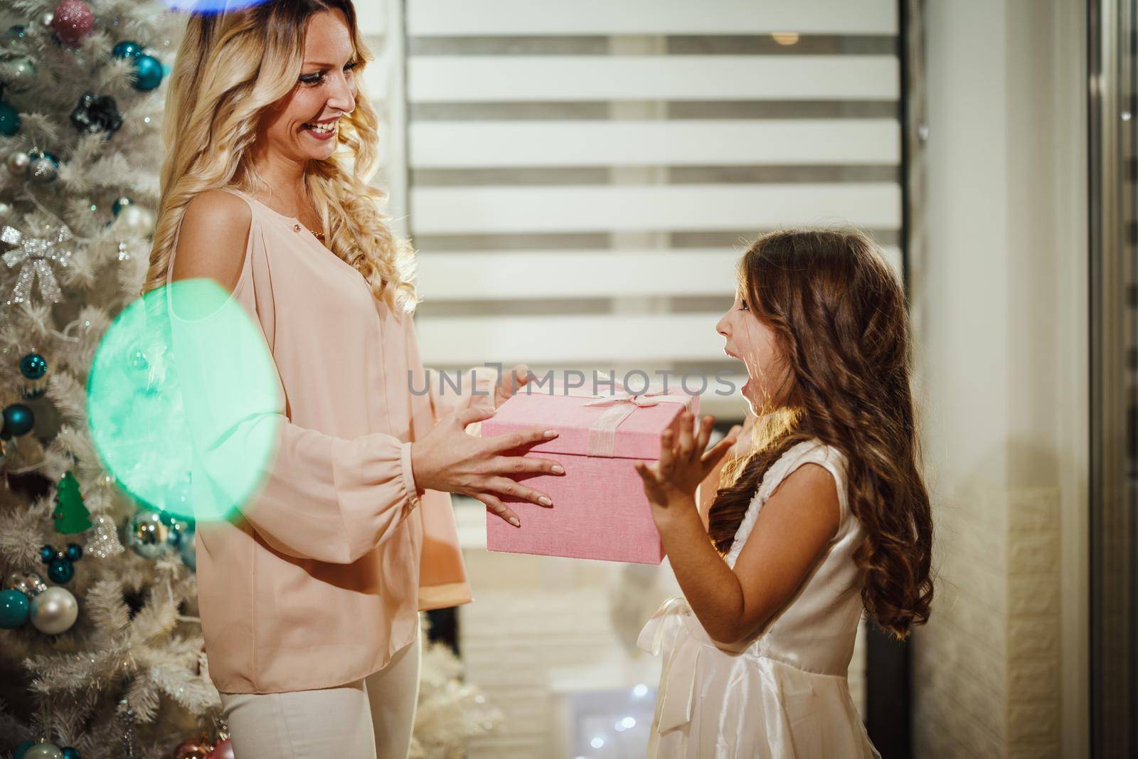 Shot of a cute little girl and her attractive mom are sitting on the floor, by the white Christmas tree and opening their Christmas presents.