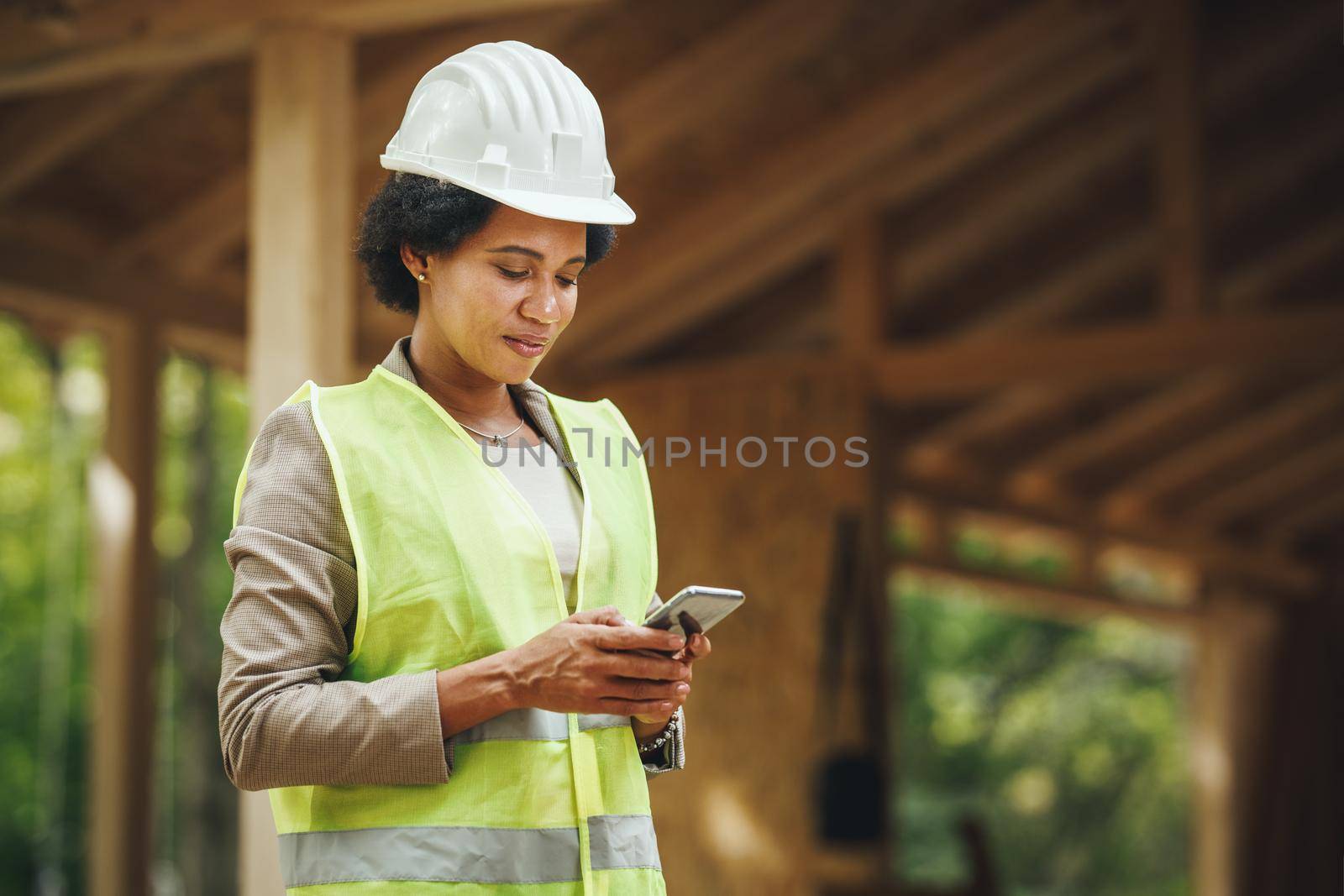 Shot of an African female architect using a smartphone and checking construction site of a new wooden house. She is wearing protective workwear and white helmet.