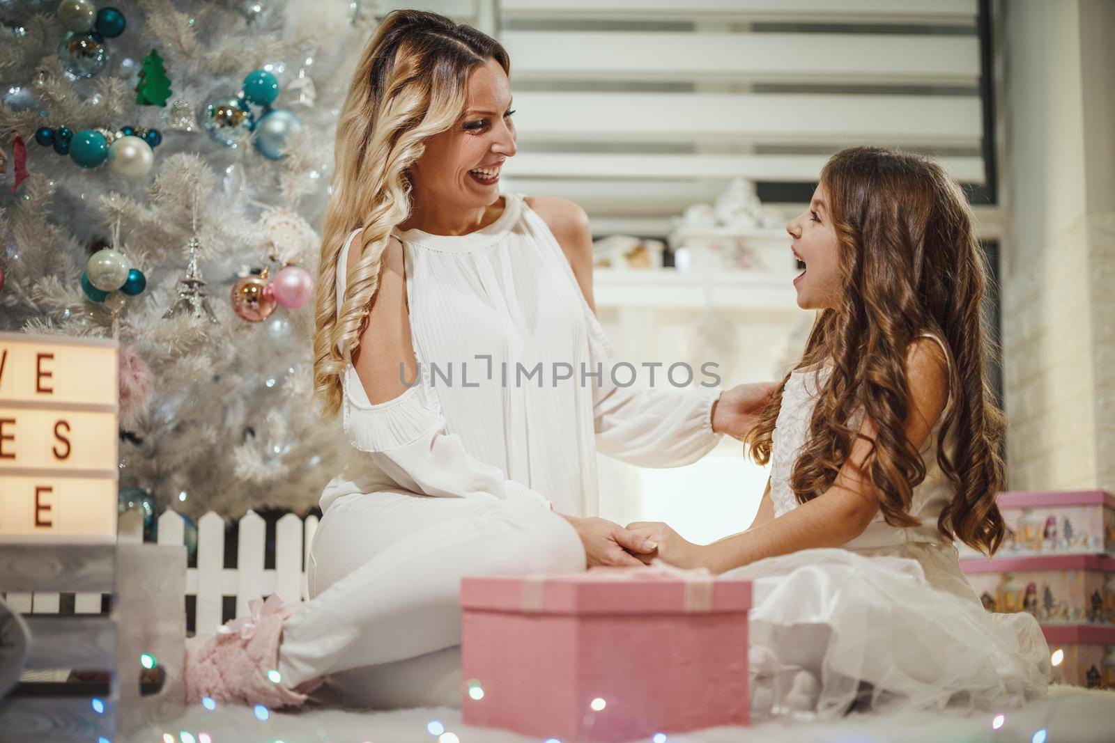 Shot of a cute little girl and her attractive mom are sitting on the floor, by the white Christmas tree and opening their Christmas presents.