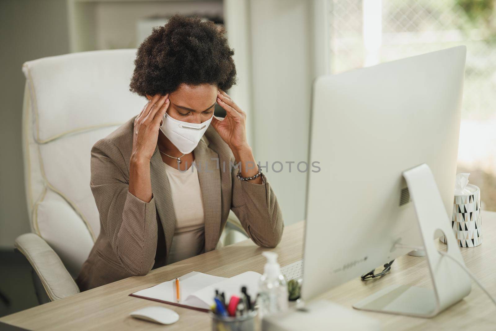 Shot of a tired African businesswoman with protective N95 mask sitting alone in her office and working on computer during COVID-19 pandemic.